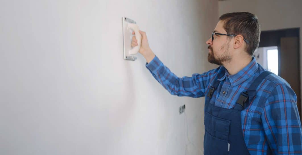 A bearded man in blue plaid shirt and overalls adjusts a white wall thermostat