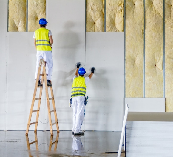 Two construction workers install drywall panels over insulation in an unfinished building interior